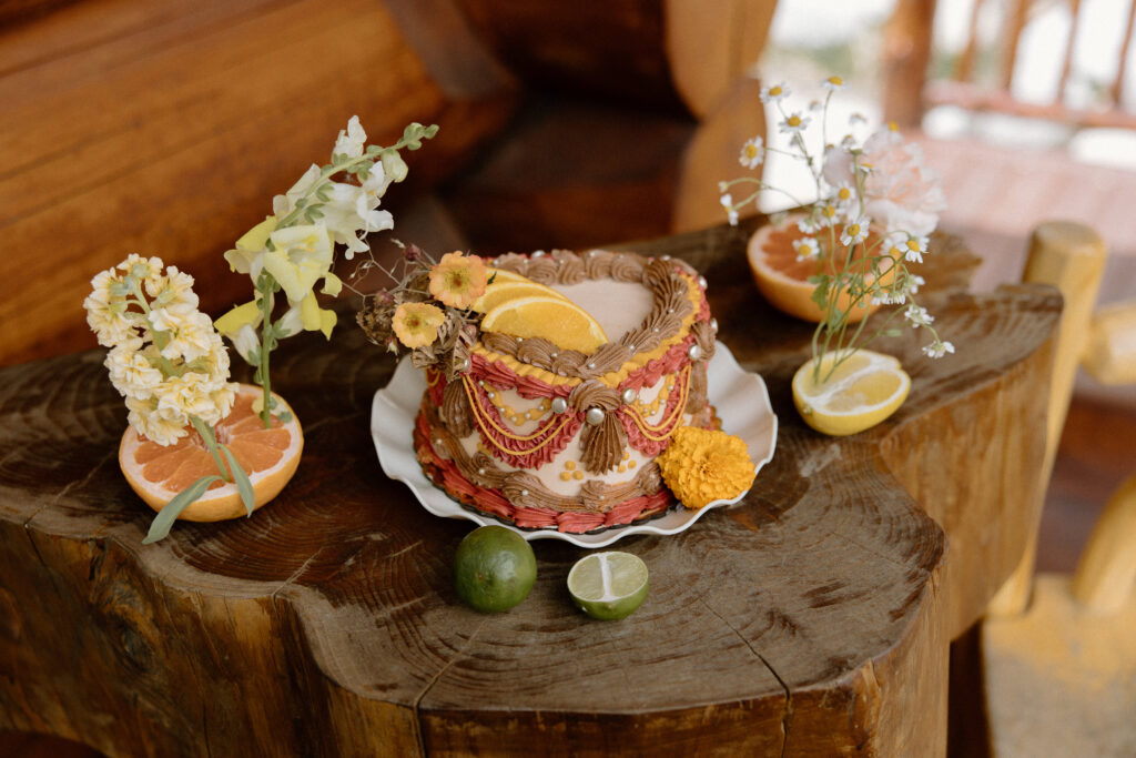 Rustic Colorado cabin elopement cake decorated with orange slices, orange florals, and layered brown, red, and white icing in a heart shape.” “Fine-art heart-shaped elopement cake featuring brown, red, and white layers topped with citrus slices and orange flowers in a cozy Colorado cabin setting.