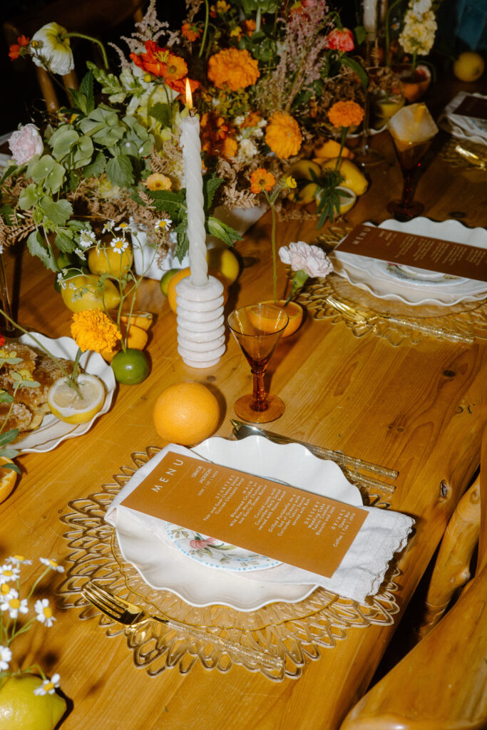 Cozy Colorado cabin elopement tablescape with orange menu, gold flatware, orange goblets, fresh citrus, orange flowers, and white candles.