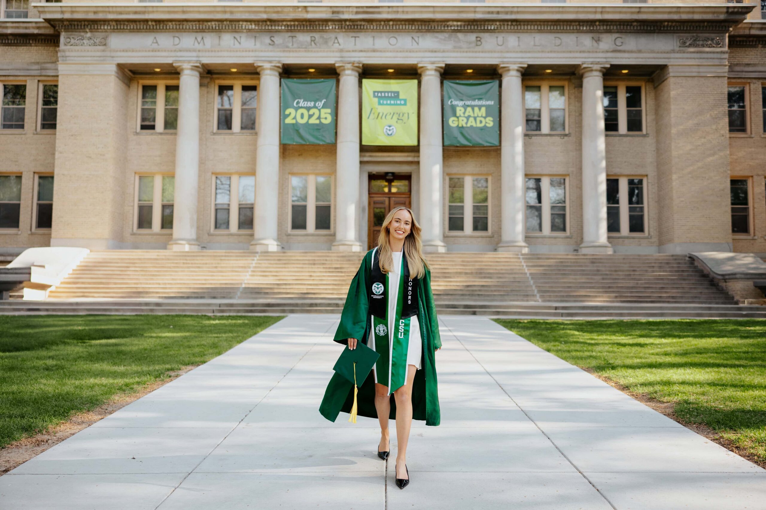 A Colorado State University graduate walking across the sidewalk in front of the Administration Building, with the large stone pillars and neoclassical architecture in the background on the CSU campus.