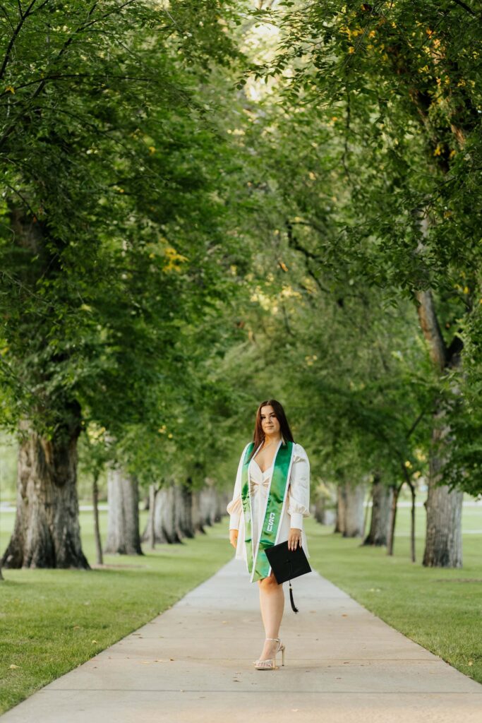 A Colorado State University graduate in a green and gold cap and gown walking down the center sidewalk of The Oval, framed by the historic American Elm trees on the CSU Fort Collins campus.