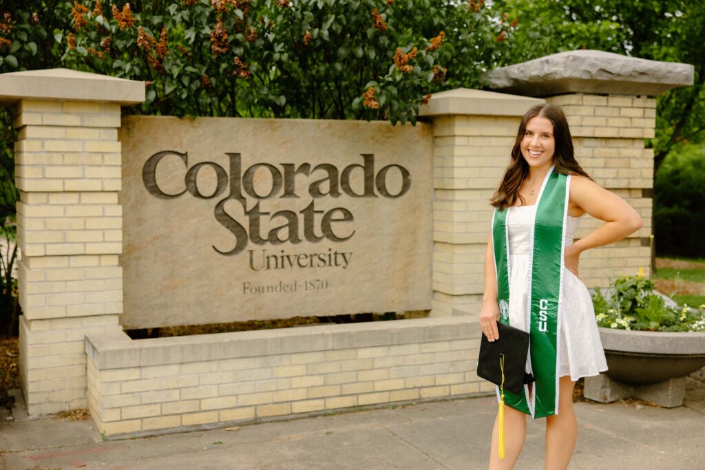 A Colorado State University graduate standing proudly in front of a brick and stone CSU monument sign on the Fort Collins campus, wearing a green and gold graduation gown.