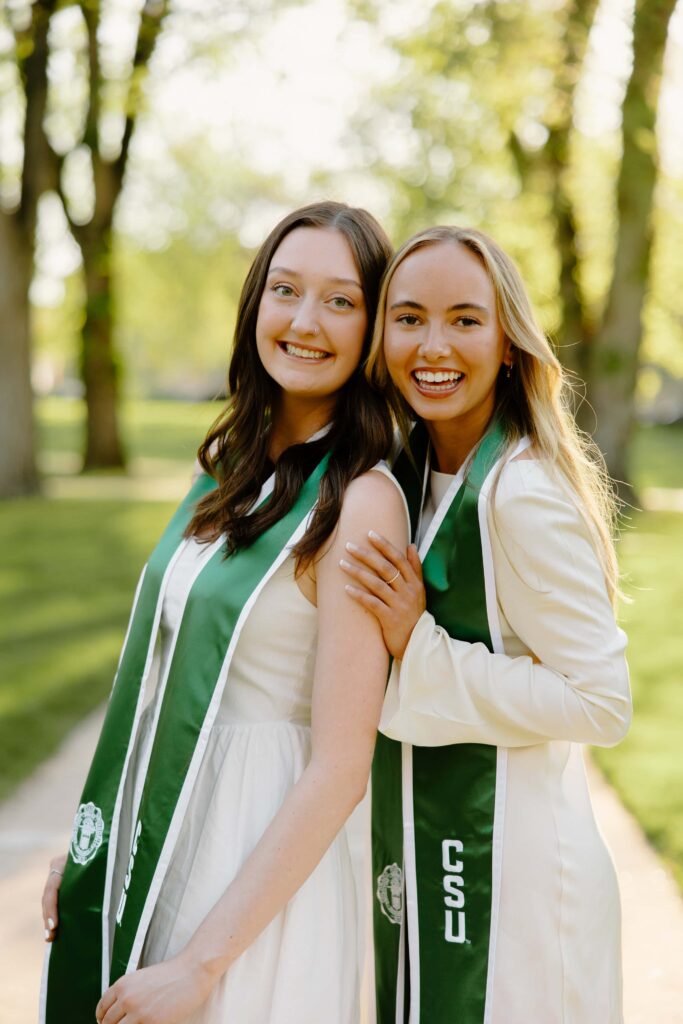 Two Colorado State University graduates in matching green and gold regalia posing together with their arms around each other on the CSU campus during commencement.