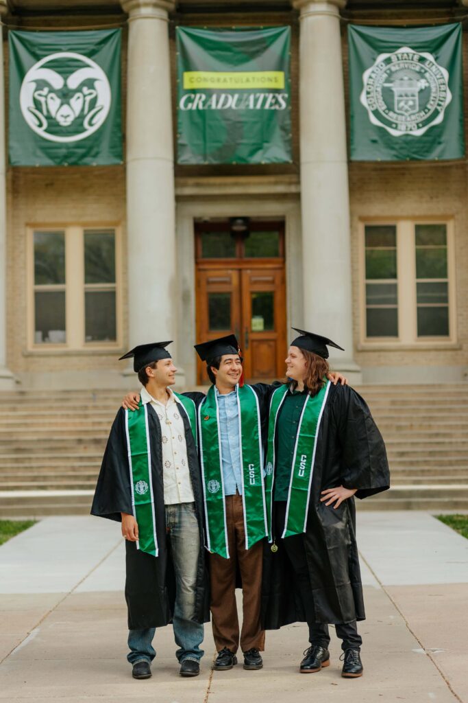 Three male Colorado State University graduates in green caps and gowns posing together in front of the historic stone pillars of the CSU Administration Building.