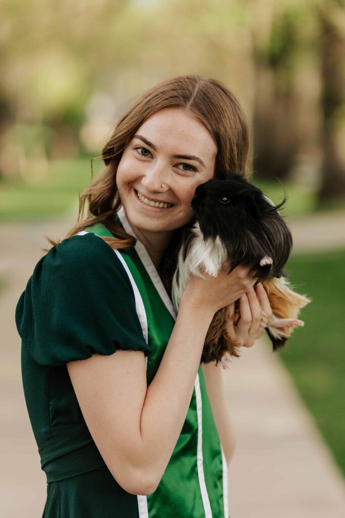A Colorado State University graduate in a cap and gown holding her pet guinea pig while posing at the historic CSU Oval in Fort Collins.