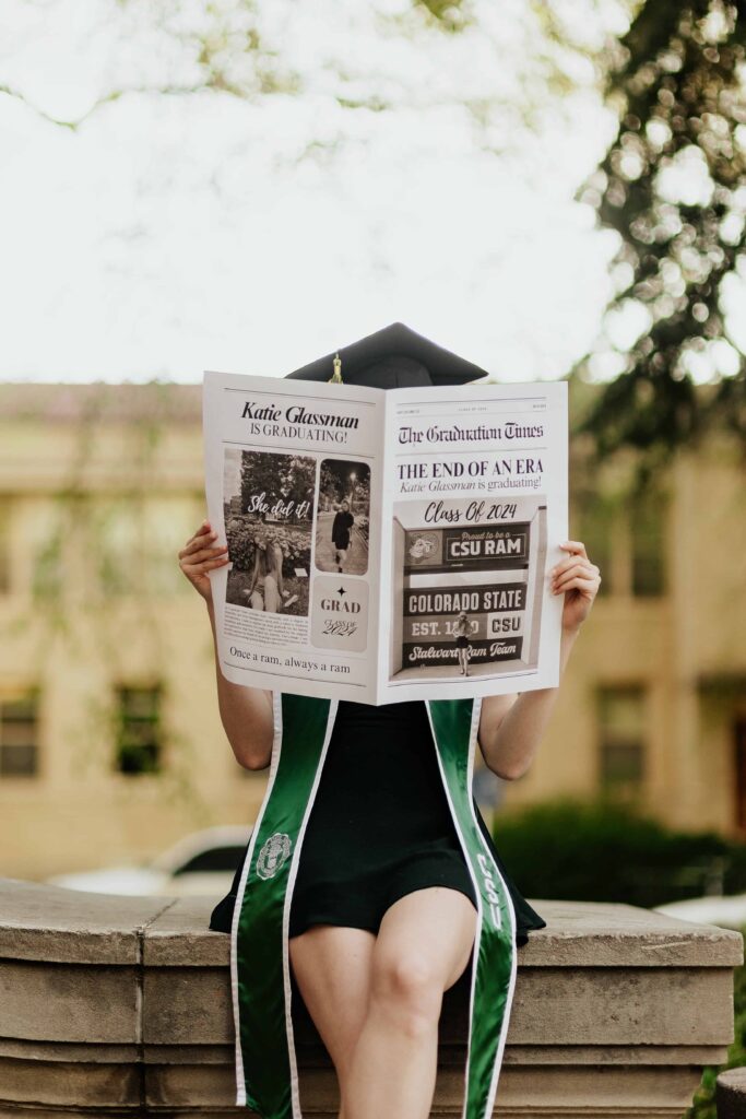 A Colorado State University graduate standing in front of the stone pillars of the CSU Administration Building while holding a newspaper to celebrate commencement in Fort Collins.