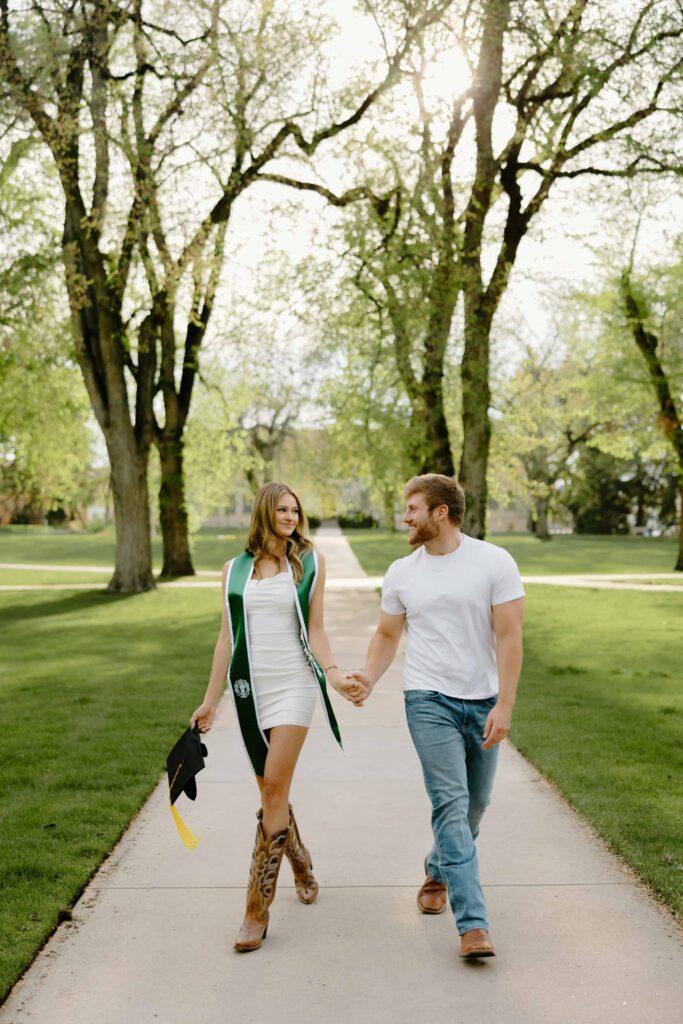 A Colorado State University graduate in a green graduation gown holding hands with her boyfriend while walking through the historic Oval on the CSU campus in Fort Collins.