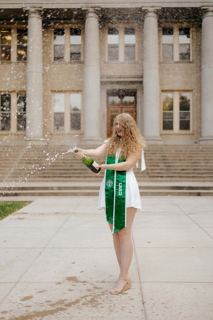 A Colorado State University graduate laughing and popping a bottle of champagne, with white foam spraying in the air, during a graduation photo session on the CSU campus.