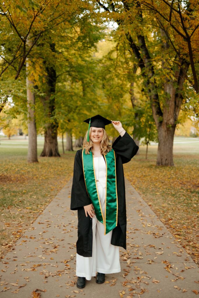 A Colorado State University graduate posing for a portrait on The Oval in the fall, surrounded by historic American Elm trees with vibrant yellow and orange autumn leaves.