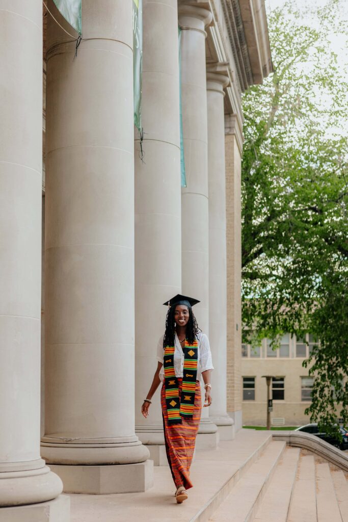 A CSU graduate smiling in front of the Colorado State University Administration Building, featuring the neoclassical architecture and wide concrete steps leading to the main entrance.