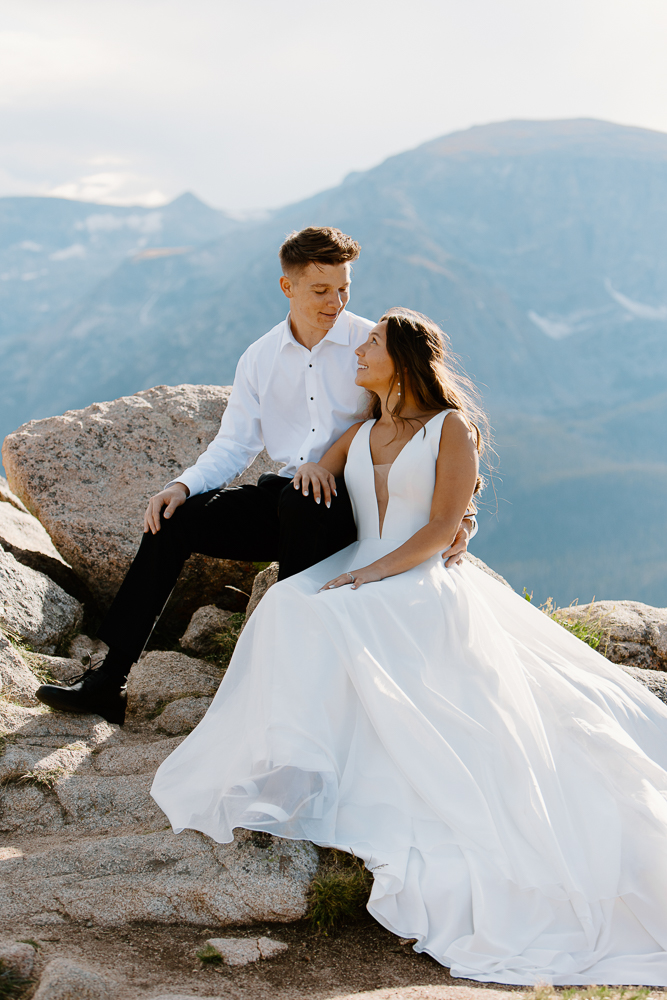 A bride and groom sit closely together on a rock along Trail Ridge Road, enjoying breathtaking views of the Colorado mountains.