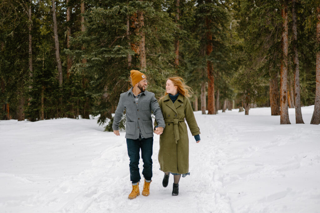 A couple runs in a forest during their Winter Engagement Photos at Echo Lake, Colorado.